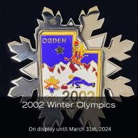 Pin in a snowflake shape with the Utah map in the center. The map showcases athletes skiing. The upper left corner of the map has the word 'Ogden' and the lower right corner says '2002'.